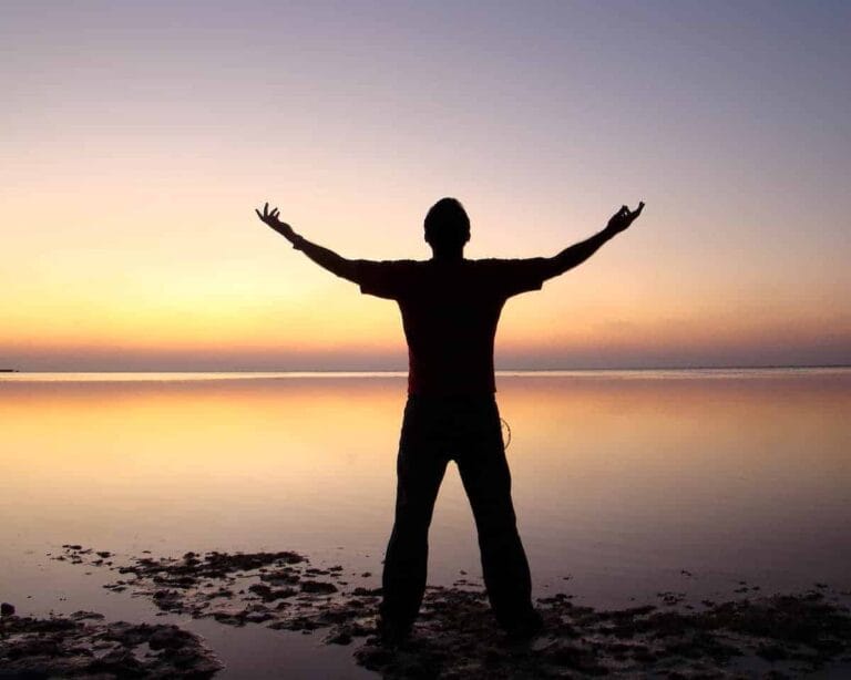 Peaceful sunset over calm water with a person standing with arms outstretched, symbolising financial growth and personal success.